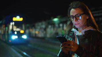 Woman stands at a transport stop at night, using smartphone and waiting for tram video