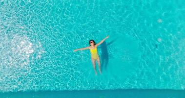 topo baixa Visão do uma mulher dentro a amarelo roupa de banho deitado em dela costas dentro a piscina. video