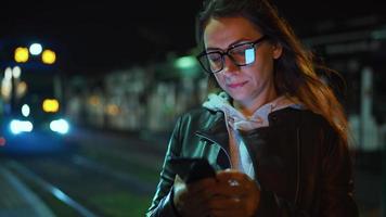 Woman stands at a transport stop at night, using smartphone and waiting for tram video