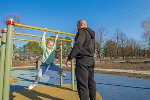 un padre ayuda su nueve años hija Halar sí misma arriba en el horizontal barras a el para niños patio de juegos. el concepto de un sano y deportivo familia, del padre día foto