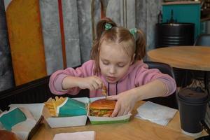 un pequeño hambriento niña con apetito come rápido comida en un café a un mesa, un hamburguesa y francés papas fritas foto