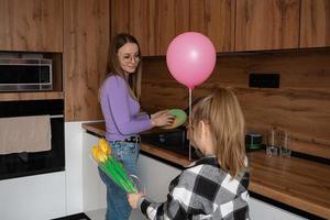 The daughter congratulates her mother on Mother's Day, gives her a balloon and flowers. The woman washes the dishes and is busy at this time with household chores. photo