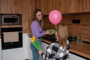 The daughter congratulates her mother on Mother's Day, gives her a balloon and flowers. The woman washes the dishes and is busy at this time with household chores. photo