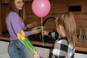 The daughter congratulates her mother on Mother's Day, gives her a balloon and flowers. The woman washes the dishes and is busy at this time with household chores.