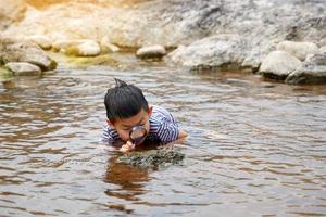 Asian boy enjoys exploring creatures in stream, along rocks with magnifying glass. concept of learning outside the classroom, natural learning resources. Soft and selective focus. photo