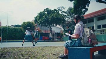 un' femmina alunno è seduta nel il scuola cortile con un' sfondo di persone giocando pallacanestro video