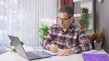 homem trabalhando dentro casa escritório estudos levando notas. homem dentro óculos trabalhando a partir de dele computador portátil às casa escritório e fazer notas em papel. video