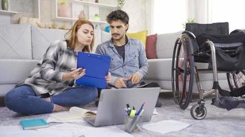 a casa, il Disabilitato uomo e il suo fidanzata siamo studiando per Lezioni. giovane uomo nel sedia a rotelle studiando con il suo fidanzata su il pavimento. video