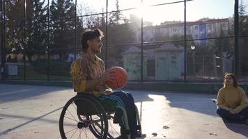 Desativado homem jogando basquetebol dentro cadeira de rodas com namorada ao ar livre. jovem homem jogando basquetebol dentro uma cadeira de rodas lança basquetebol e regozija-se com dele namorada. video