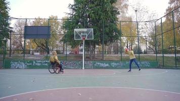 gehandicapt jong Mens Toneelstukken basketbal met zijn vriendin Aan een buitenshuis basketbal rechtbank. de meisje wie helpt de gehandicapt tiener Speel basketbal is gelukkig. video