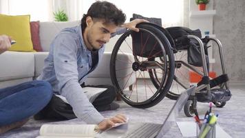 Disabilitato adolescente utilizzando un' sedia a rotelle è studiando a casa. Disabilitato giovane uomo studiando a casa con il suo fidanzata, guardare a libri e assunzione Appunti. video