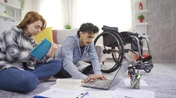 A student with a disability and a young girl look at a laptop and books at home. Disabled teenager using a wheelchair is studying at home with his girlfriend. video