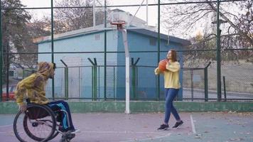 gehandicapt jong Mens in rolstoel spelen basketbal met zijn vriendin. gehandicapt jong Mens en zijn vriendin spelen basketbal buiten in langzaam beweging. video