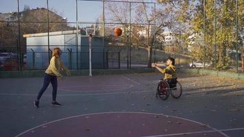 jovem homem dentro cadeira de rodas e dele namorada jogando basquetebol dentro lento movimento. Desativado jovem homem tocam basquetebol com dele namorada em a ao ar livre basquetebol tribunal. video