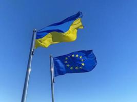 saludó ucranio y europeo Unión banderas en astas de bandera en contra azul cielo foto