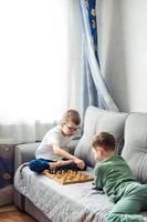 Boys playing wooden chess lying on a gray sofa in front of the window photo