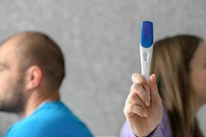 un hombre y un mujer son sentado en el cama espalda a atrás, ellos peleado, el hombre es no Listo y lo hace no querer niños, el mujer tiene un positivo el embarazo prueba en su manos. foto