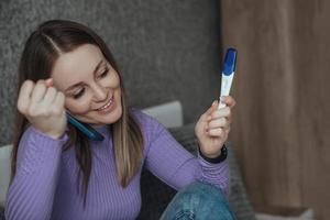 un mujer es sentado en un cama, en su manos es un teléfono y un negativo el embarazo prueba, ella es contento. sin hijos mujer, consciente rechazo de maternidad foto