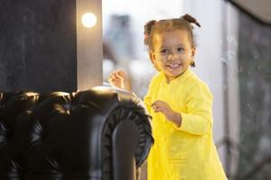 un pequeño africano americano niña soportes cerca el espejo y sonrisas foto