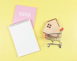 flat layout of wooden house model in shopping trolley or shopping cart  with   blank page notebook and pink diary or  2023   on yellow  background with copy space, home purchase plan  concept.