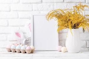 Pastel colored Easter eggs and mimosa flowers with blank white frame for mockup design, front view on white brick wall background photo