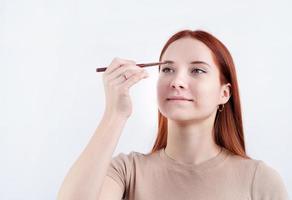 alegre pelirrojo joven mujer utilizando maquillaje cepillo haciendo arriba aislado en blanco antecedentes foto