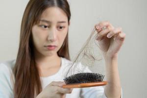 Serious, worried asian young woman, girl holding brush, show her comb, hairbrush with long loss hair problem after brushing, hair fall out on her hand in living room. Health care, beauty treatment. photo