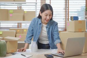 Small business entrepreneur SME, asian young woman owner packing product, checking parcel box delivery, using laptop receive order from customer, working at home office. Merchant online, ecommerce. photo