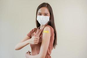 Portrait of health care asian young woman wearing face mask protect, getting vaccine of covid-19, influenza in casual, showing arm on yellow bandage, plaster, isolated on nature background, copy space