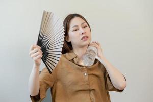 Summer heat stroke, hot weather, tired asian young woman sweaty and thirsty, refreshing with hand in blowing, wave fan to ventilation, holding cold water bottle tap her body when temperature high. photo