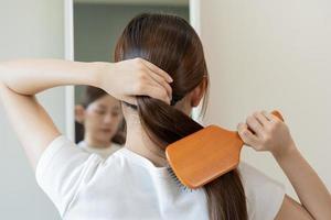 Health hair care, beauty makeup asian woman, girl holding hairbrush and brushing, combing her long straight hair looking at reflection in mirror in morning routine after salon treatment, hairstyle. photo