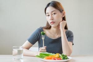 dieta en aburrido, infeliz hermosa asiático joven mujer, niña en haciendo dieta, participación tenedor a brócoli en ensalada lámina, disgusto o cansado con comer Fresco vegetales. nutrición de limpio, sano comida bueno gusto. foto