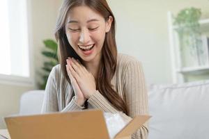 contento entusiasmado, asiático joven mujer, niña cliente sentado en sofá a hogar, apertura y desempacar cartulina caja caja de cartón paquete o empaquetar después comprando ordenando presente, compras en línea, entrega Servicio concepto. foto