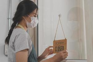 Asian young owner retail,coffee shop woman turning sign board to closed with wearing face mask,protection to pandemic of coronavirus.Close store,restaurant or pub due to lockdown, quarantine of covid. photo