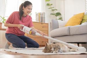 Happy asian young housekeeper woman using vacuum cleaning, cleaner to remove dust, hair or fur on floor in living room while cute cat lying on carpet. Routine housework, chore in household of maid. photo