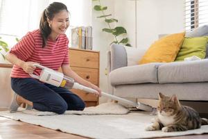 contento asiático joven ama de casa mujer utilizando vacío limpieza, limpiador a eliminar polvo, pelo o piel en piso en vivo habitación mientras linda gato acostado en alfombra. rutina tareas del hogar, faena en casa de mucama. foto