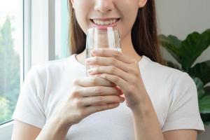 Happy beautiful, asian young woman, girl drinking, sip fresh glass of water for hydration of body, holding transparent glass in her hand, thirsty at home. Health care, healthy lifestyle concept. photo