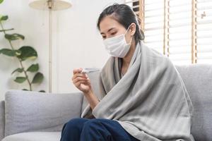 enfermo, influenza asiático joven mujer, niña vistiendo cara máscara en dolor de cabeza tener un fiebre, gripe y cheque termómetro medida cuerpo temperatura, sensación enfermedad sentado en sofá cama a hogar. salud cuidado, COVID-19. foto