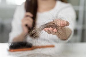 Serious, worried asian young woman, girl holding brush, show her comb, hairbrush with long loss hair problem after brushing, hair fall out on her hand in living room. Health care, beauty treatment. photo