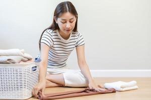 Housewife, asian young woman hand in many folding freshly shirts, sweaters or dress on desk, table after washing clean clothes and drying, making household working in room at home. Laundry and maid. photo