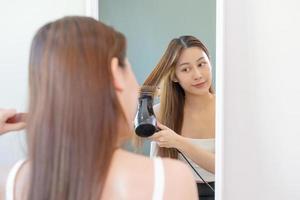 pelo secadora, feliz, asiático joven mujer, niña mirando dentro espejo reflejar utilizando secador de pelo a seco, soplo largo rubia Derecho después ducha a hogar. peluquería, pelo tratamiento y belleza salud cuidado. foto