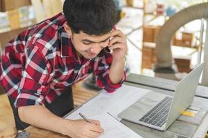 propietario empresario puesta en marcha pequeño tienda negocio. carpintero asiático joven hombre utilizando teléfono inteligente, móvil hablar con cliente acerca de orden, utilizar ordenador portátil computadora para márketing en línea. fábrica industria de madera. foto