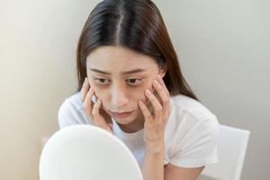 Bored, insomnia asian young woman, girl looking at mirror, without makeup, touch under eyes with problem of dark circles, puffiness, swollen or wrinkles on face. Sleepless, sleepy people, copy space photo