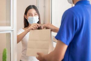 Smile, Happy asian young woman or girl  wearing face mask protect to pandemic of covid-19 and received paper bag from delivery man, messenger in uniform while lockdown, quarantine at front door home. photo