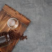 Photography of coffee beans spilling from a jar, on a piece of wood with a gray background, suitable for photos of food and beverage products, with a square photo format