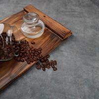 Photography of coffee beans spilling from a jar, on pieces of wood with a gray background, suitable for photos of food and beverage products, with perspective angle square photo format