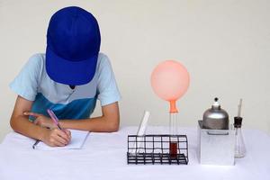 joven chico estudiante usa azul gorra , escribir en papel, hacer Ciencias experimentar acerca de globo. concepto, acción investigación. Ciencias proyecto trabajar. aprendizaje por haciendo y observando educación. yo estudiar. foto