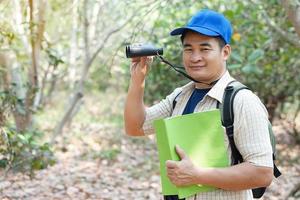 Asian man explorer wears blue cap, holds binocular in forest to survey botanical plants and creatures wildlife. Concept, nature exploration. Ecology and Environment. photo