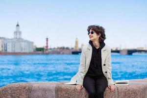 Caucasian happy girl sits on granite curb city with book. In a white coat photo