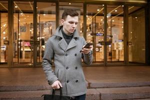 Smiling young man in gray coat is holding scroll text messages in his mobile photo